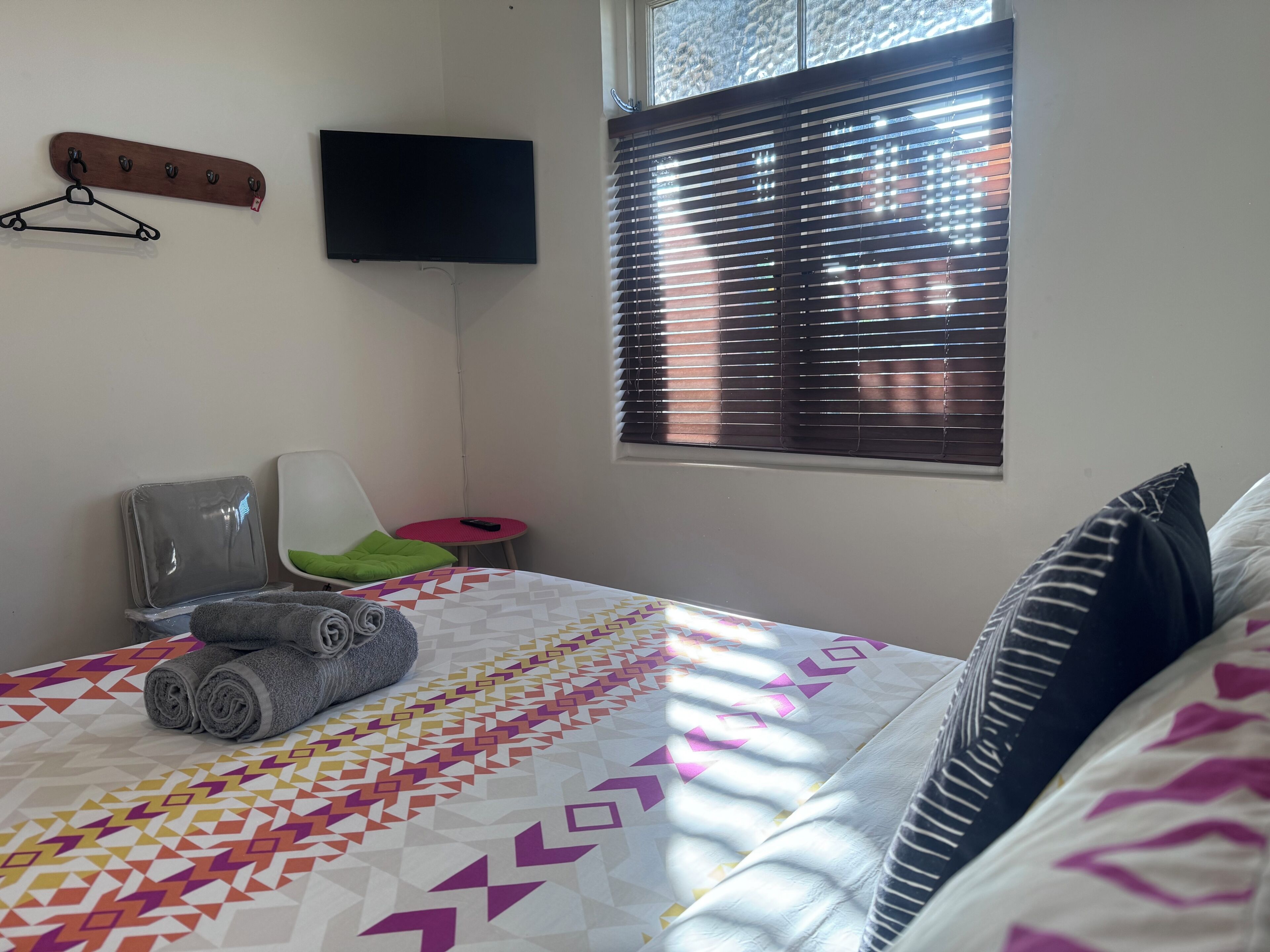 Un dormitorio con cama, una silla, televisor y ventana con persianas.