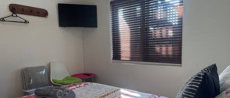 Un dormitorio con cama, una silla, televisor y ventana con persianas.