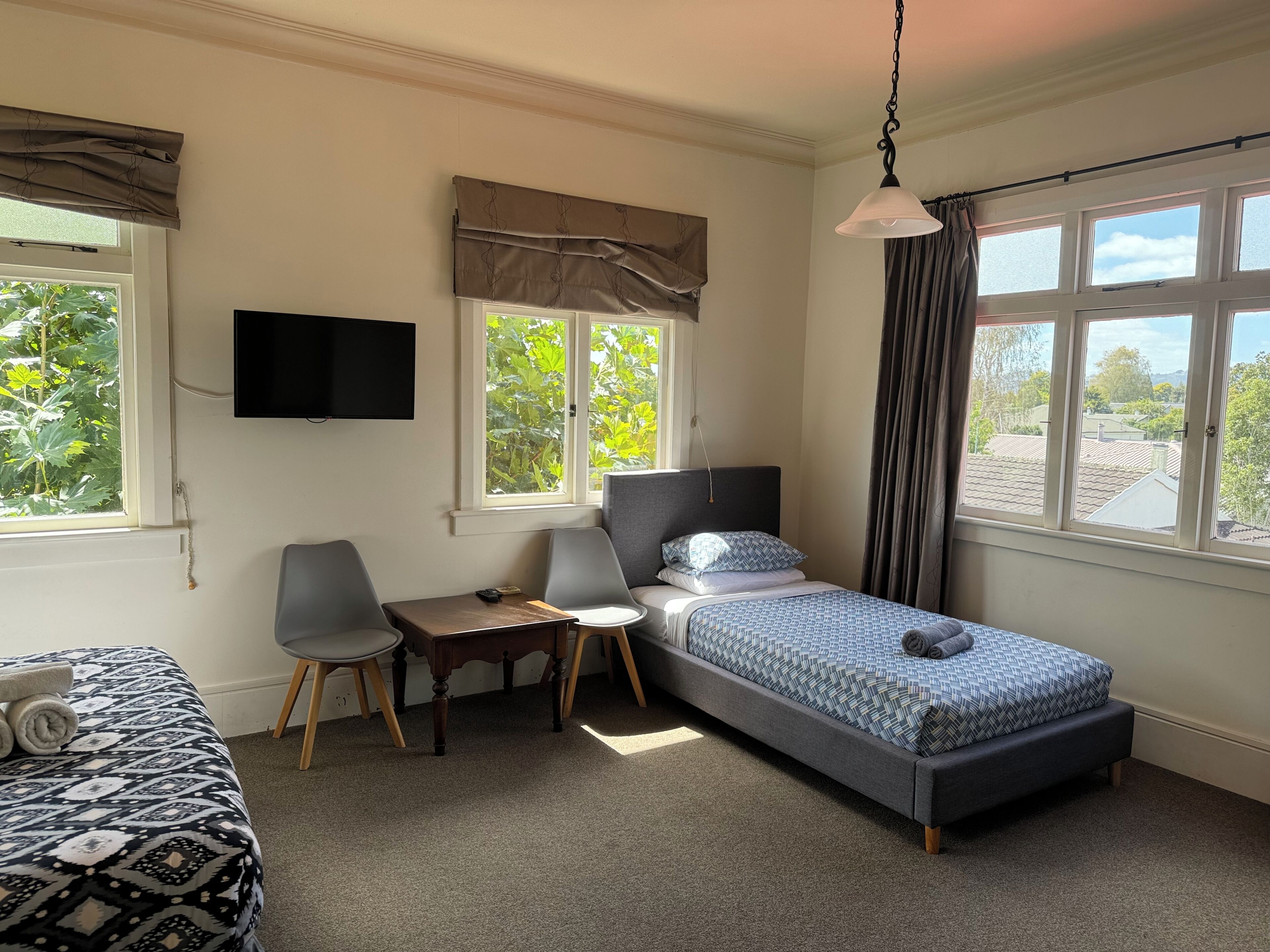 Quarto com cama, escrivaninha, cadeira, televisão e janelas com cortinas.