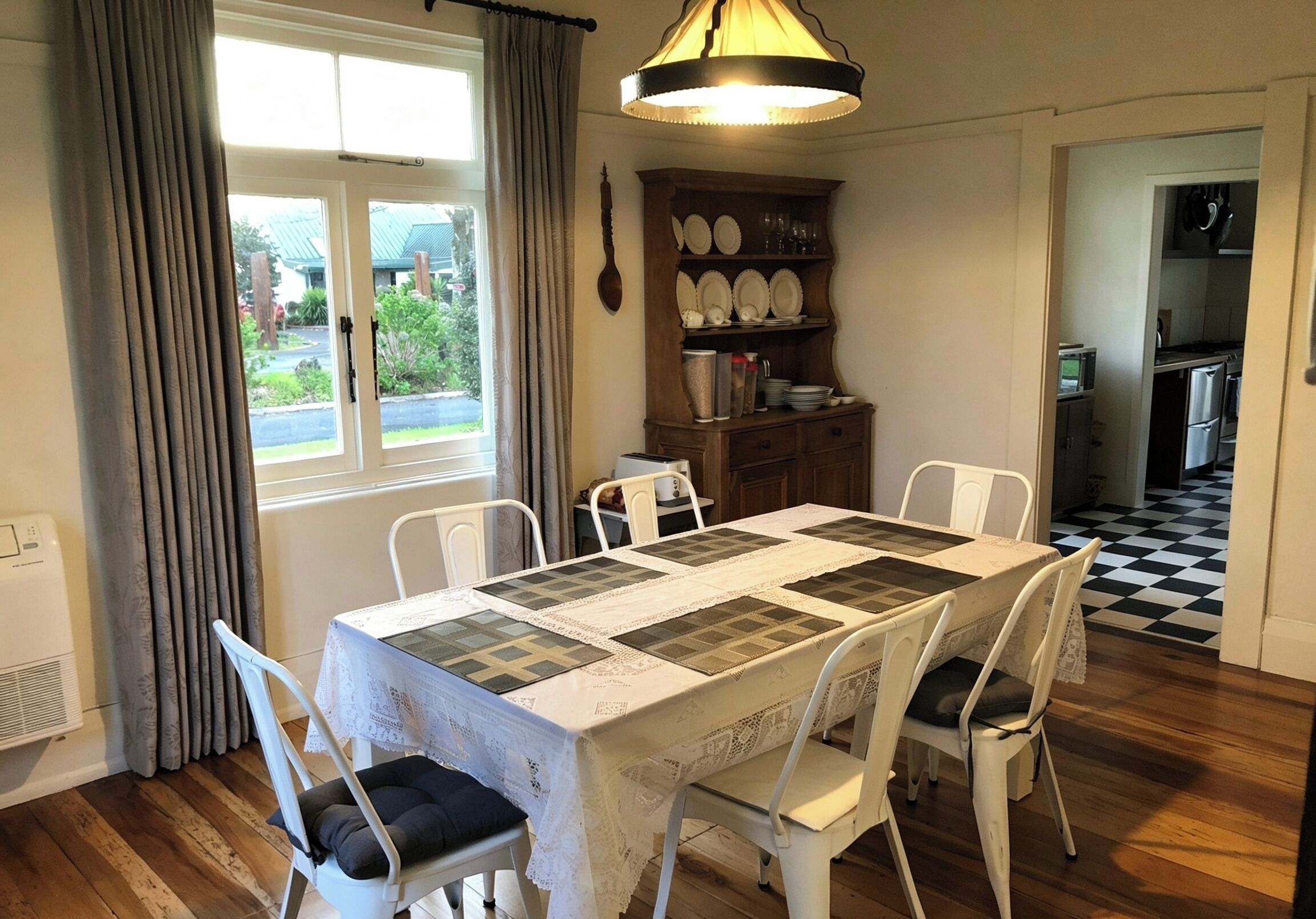 Un comedor con una mesa dispuesta para cuatro, sillas blancas, una ventana con cortinas, un mueble de madera con vajilla, y una cocina visible a través de una puerta abierta.