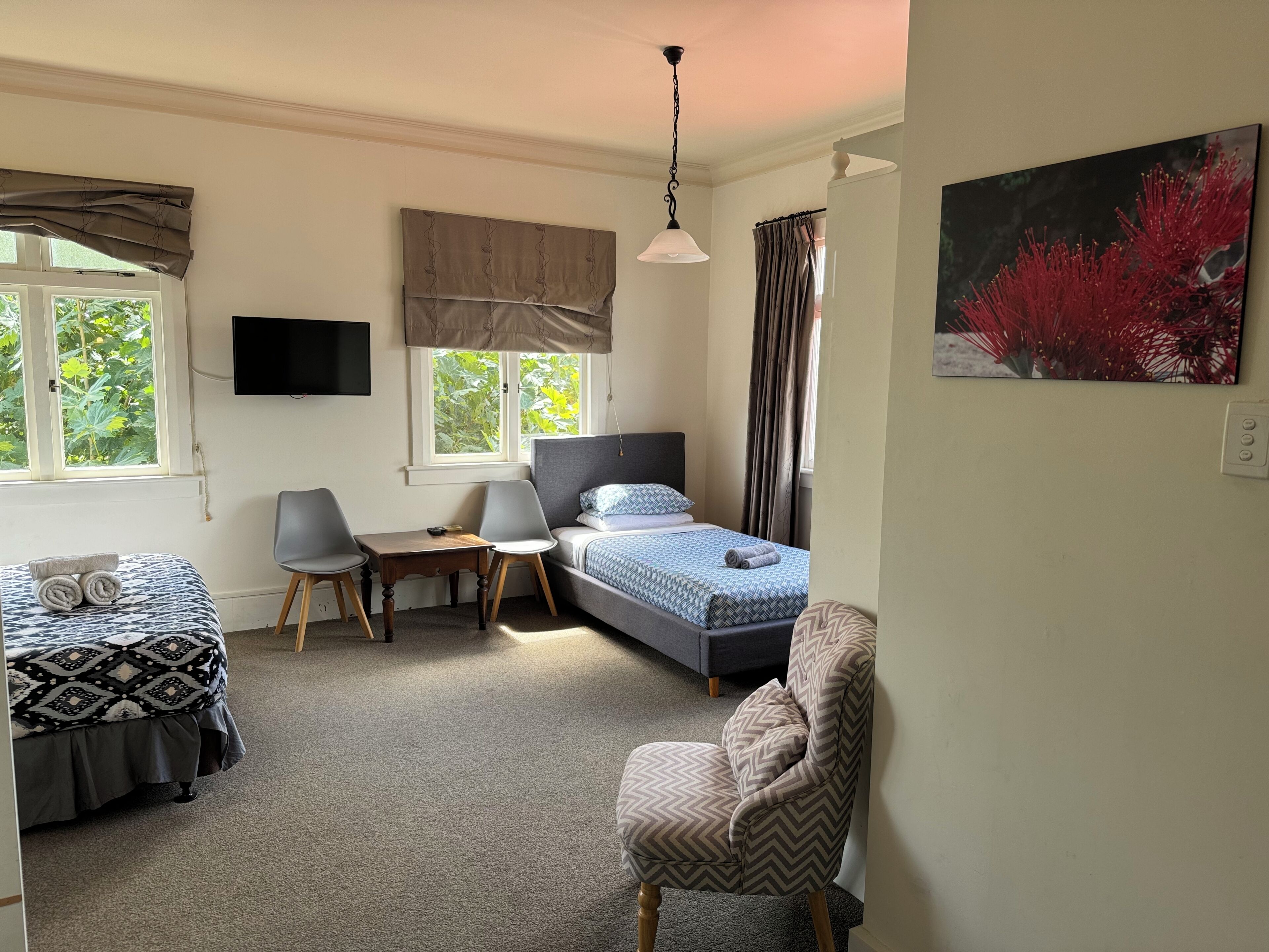 Ein Schlafzimmer mit einem Bett, zwei Stühlen, einem Schreibtisch, einem Fernseher und einem Fenster mit Vorhängen.
