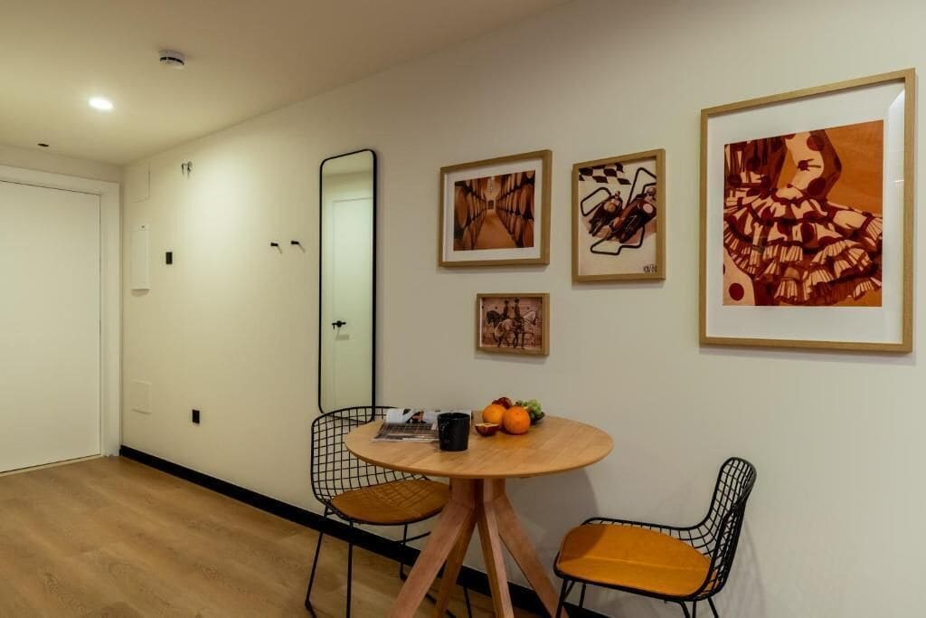 Una habitación con una mesa redonda de madera, dos sillas de alambre, un espejo y tres cuadros en la pared.