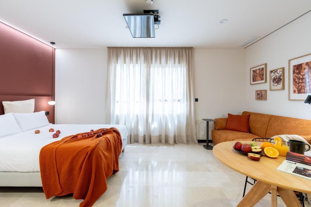 Une chambre d’hôtel avec un lit, un canapé, une table à manger avec des fruits et une fenêtre avec des rideaux.