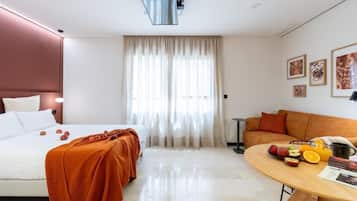 Une chambre d’hôtel avec un lit, un canapé, une table à manger avec des fruits et une fenêtre avec des rideaux.