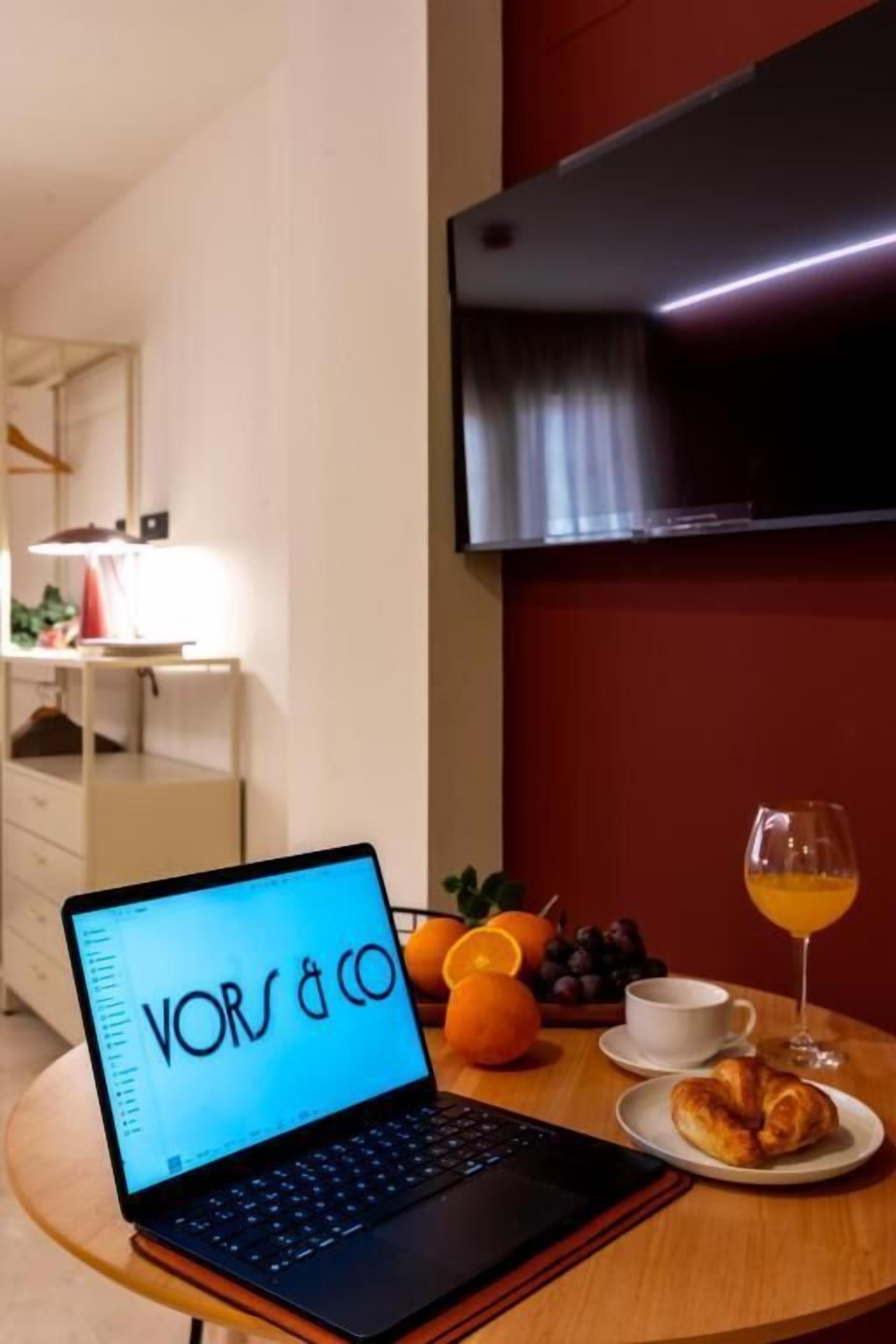 A laptop on a table with a glass of orange juice and a croissant, a white cup, and a bowl of fruit.