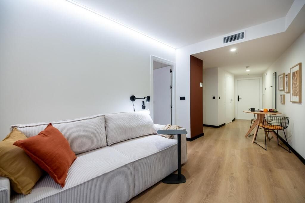 A modern living room with a sofa, a small table, and a chair. There is a door leading to another room and a hallway with a door at the end.