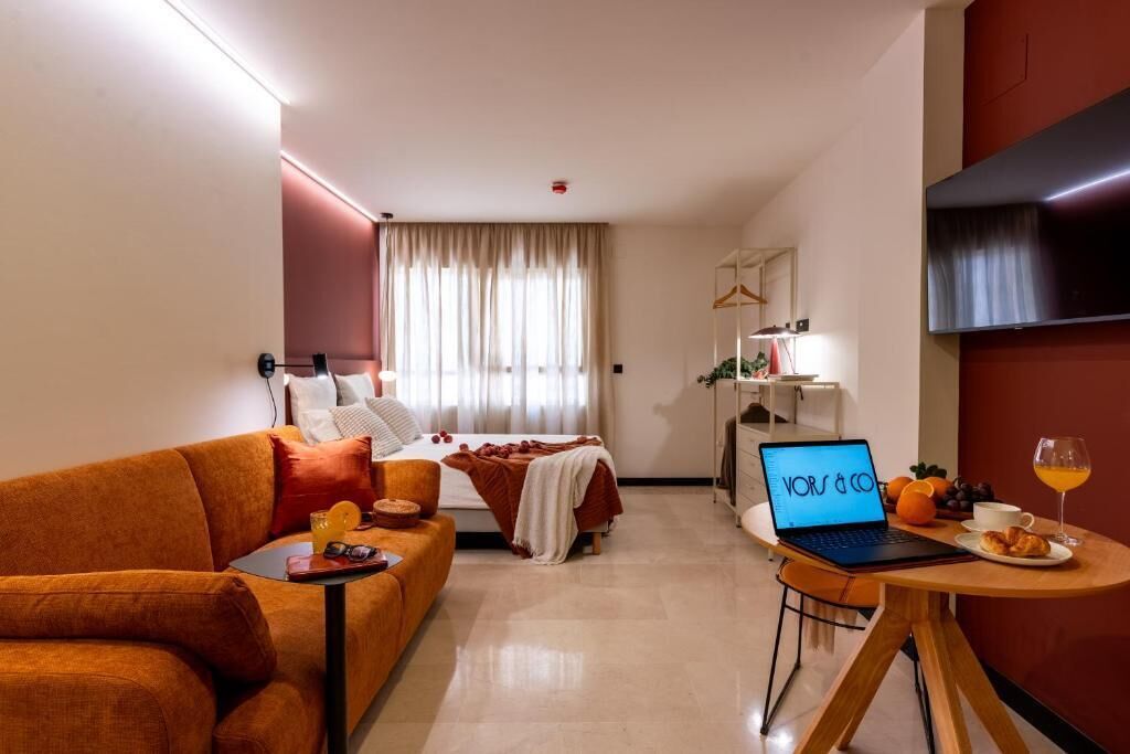 Una habitación de hotel moderna con cama, sofá, escritorio con computadora portátil, mesa de comedor con alimentos para el desayuno y un televisor montado en la pared.