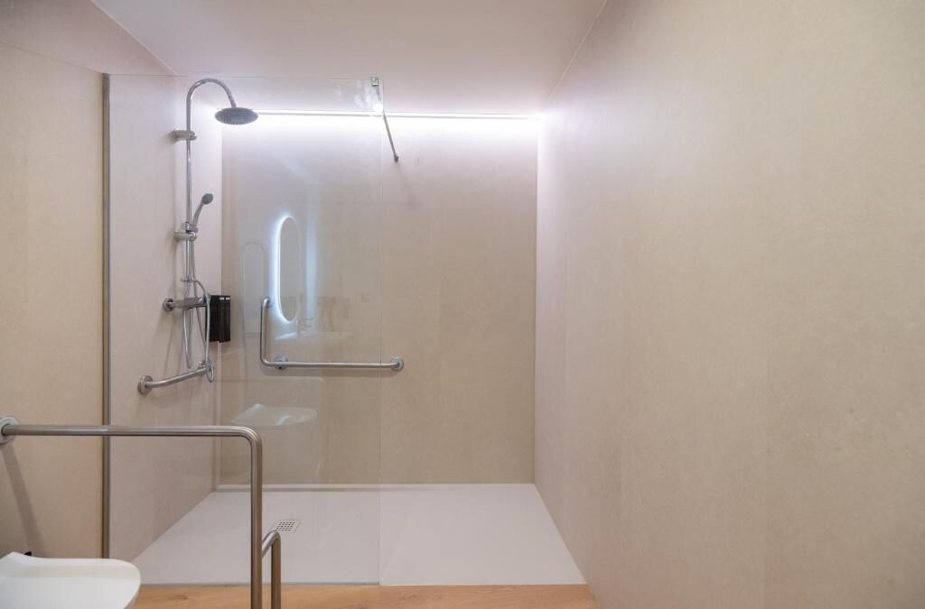 A modern bathroom with a glass shower enclosure, a handrail, and a white sink.