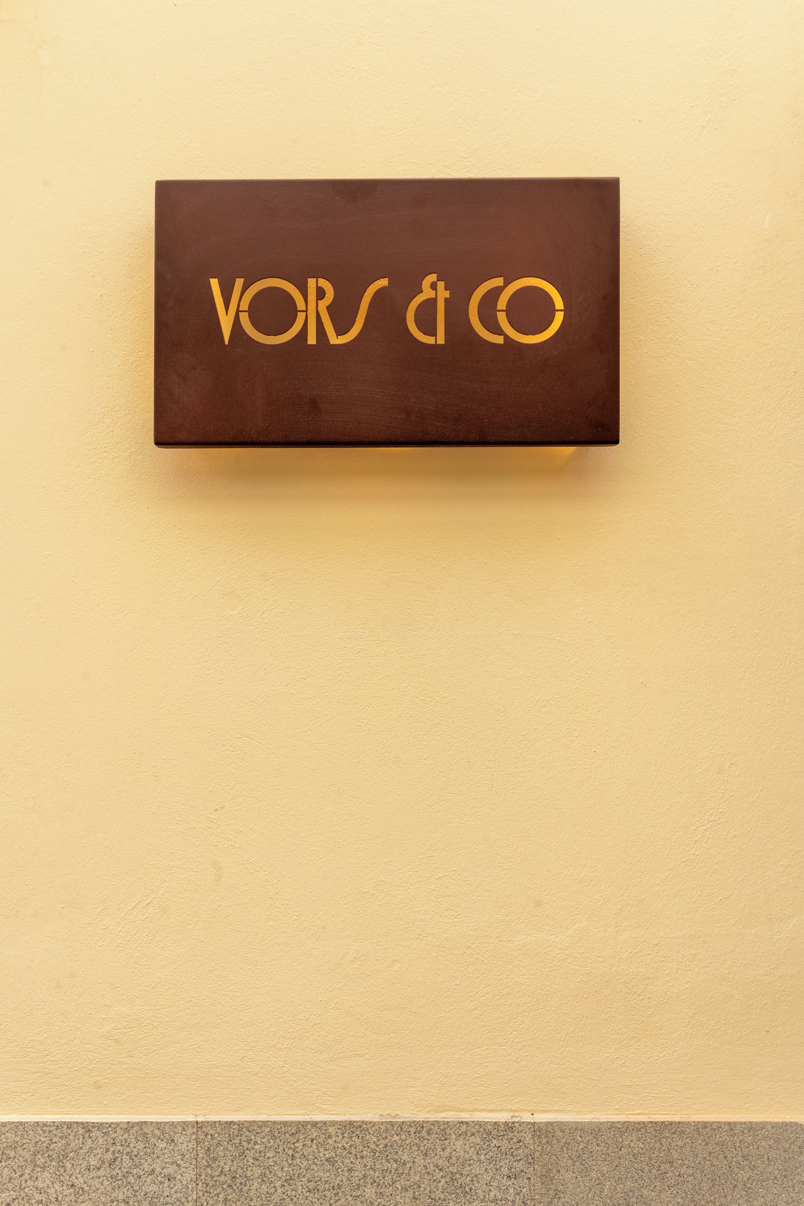 Ein braunes, rechteckiges Schild mit goldener Schrift auf einer beigen Wand.