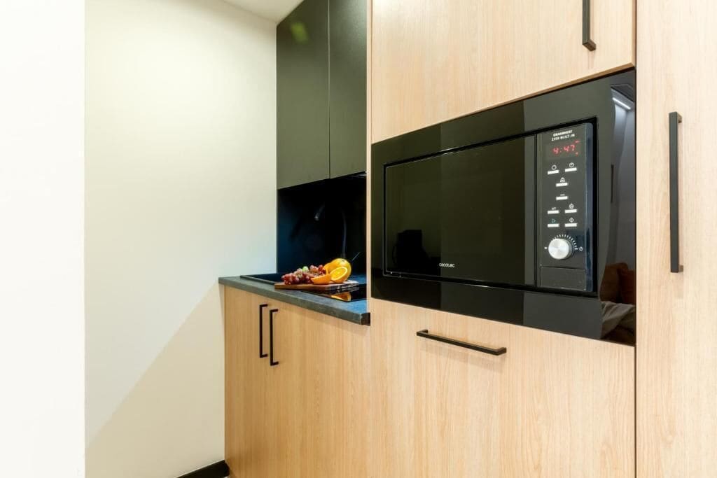 Una cucina con un forno a microonde, una sveglia digitale e una ciotola di frutta sul piano di lavoro.