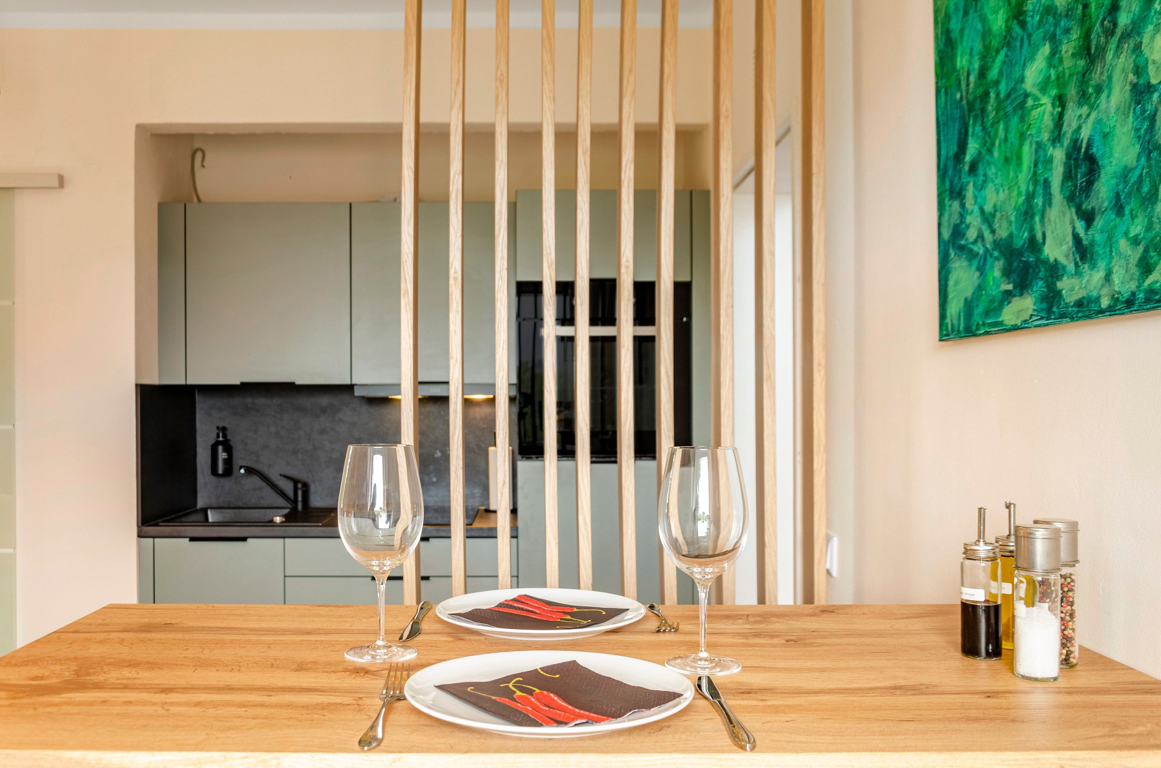 A dining table with two plates, two wine glasses, and a set of cutlery. There is a kitchen in the background with cabinets and a countertop.