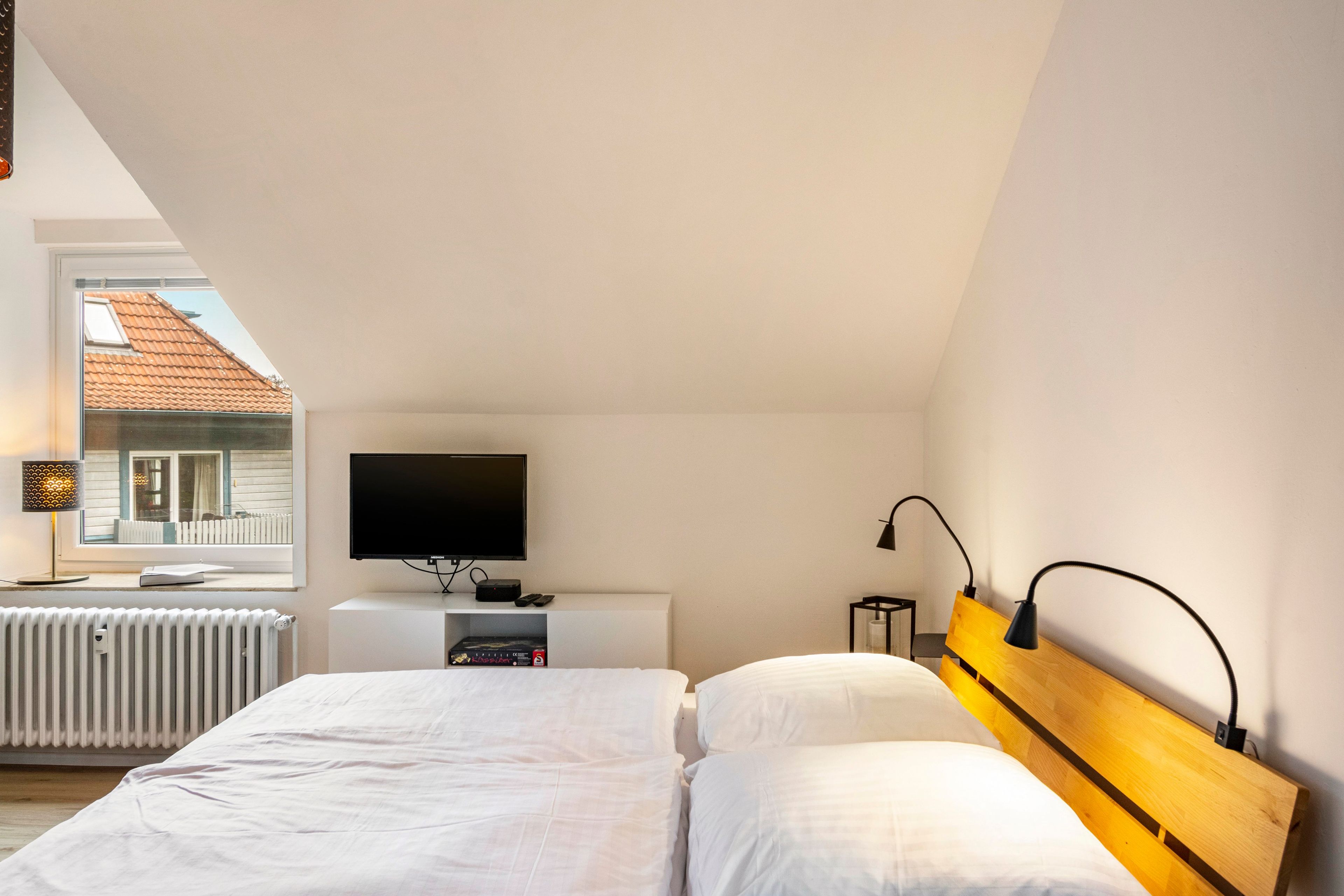 Un dormitorio con cama, televisor, mesita de noche y ventana con vista a una casa.