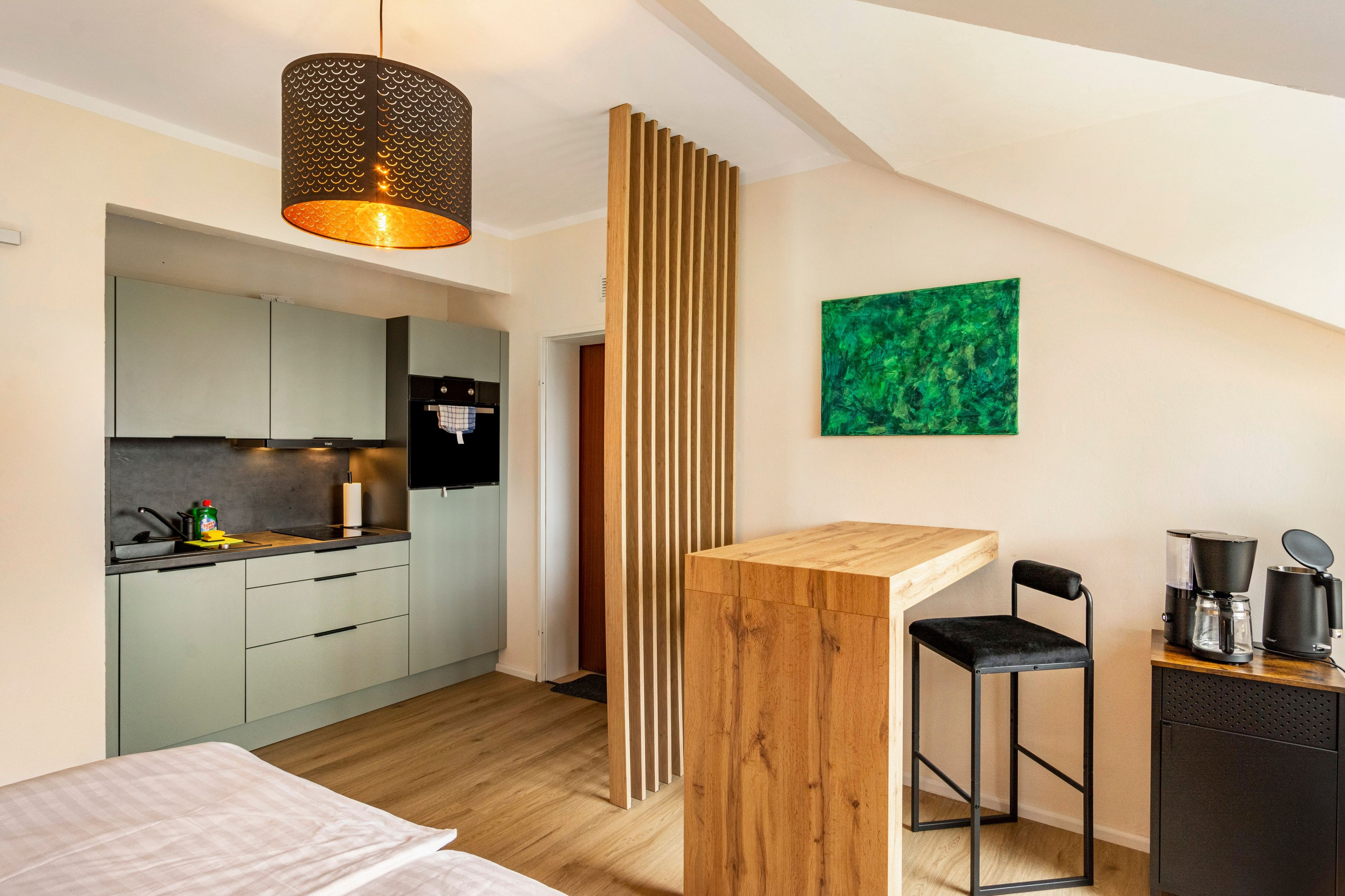 A compact room with a kitchenette, a wooden partition, a bed, a small table with a chair, and a green abstract painting on the wall.