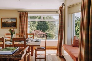 Ein Essbereich mit Tisch für zwei Personen, einem Fenster mit Vorhängen, einem Bücherregal mit Büchern und Blick ins Grüne.