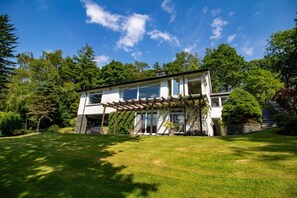 Een twee verdiepingen tellend huis met een overdekte veranda, omringd door een verzorgd gazon en bomen.