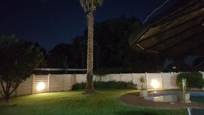 Un espacio exterior nocturno con una palmera, una estructura cubierta y una valla bien iluminada.