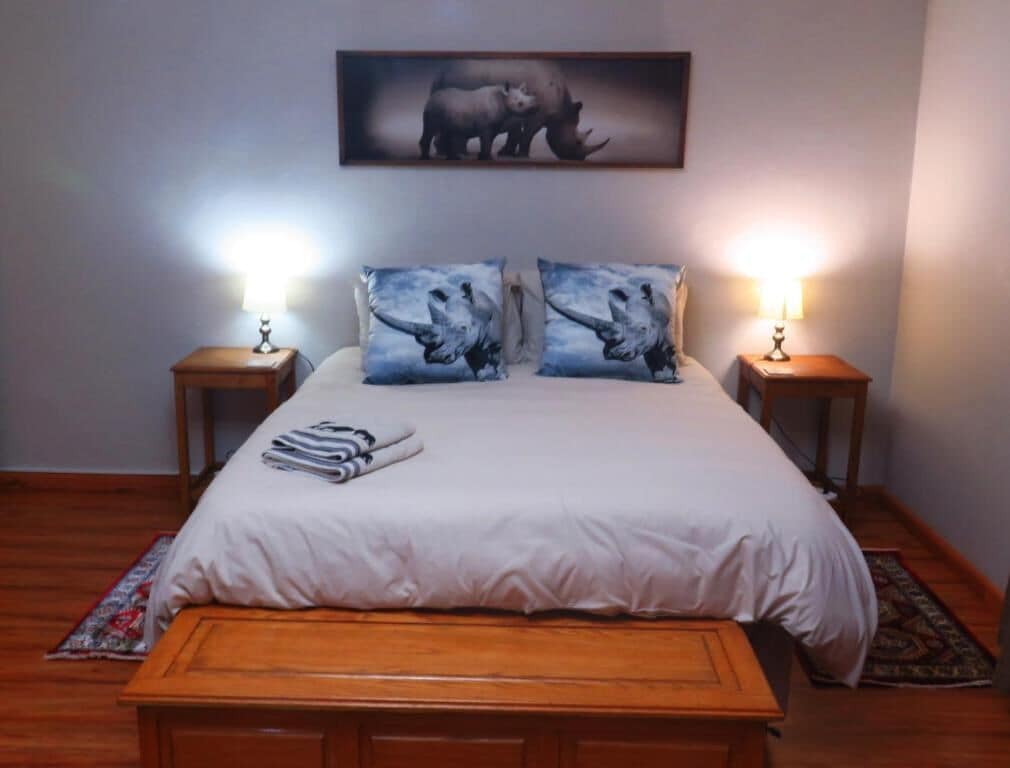 Quarto com cama grande, duas luminárias de cabeceira, um banquinho de madeira e um quadro na parede.
