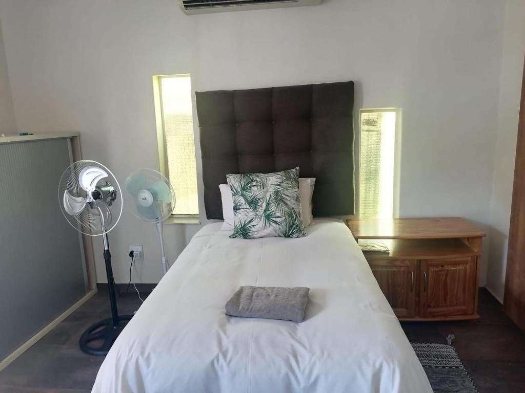 A bedroom with a bed, bedside tables, a standing fan, and a window with blinds.