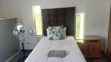 A bedroom with a bed, bedside tables, a standing fan, and a window with blinds.