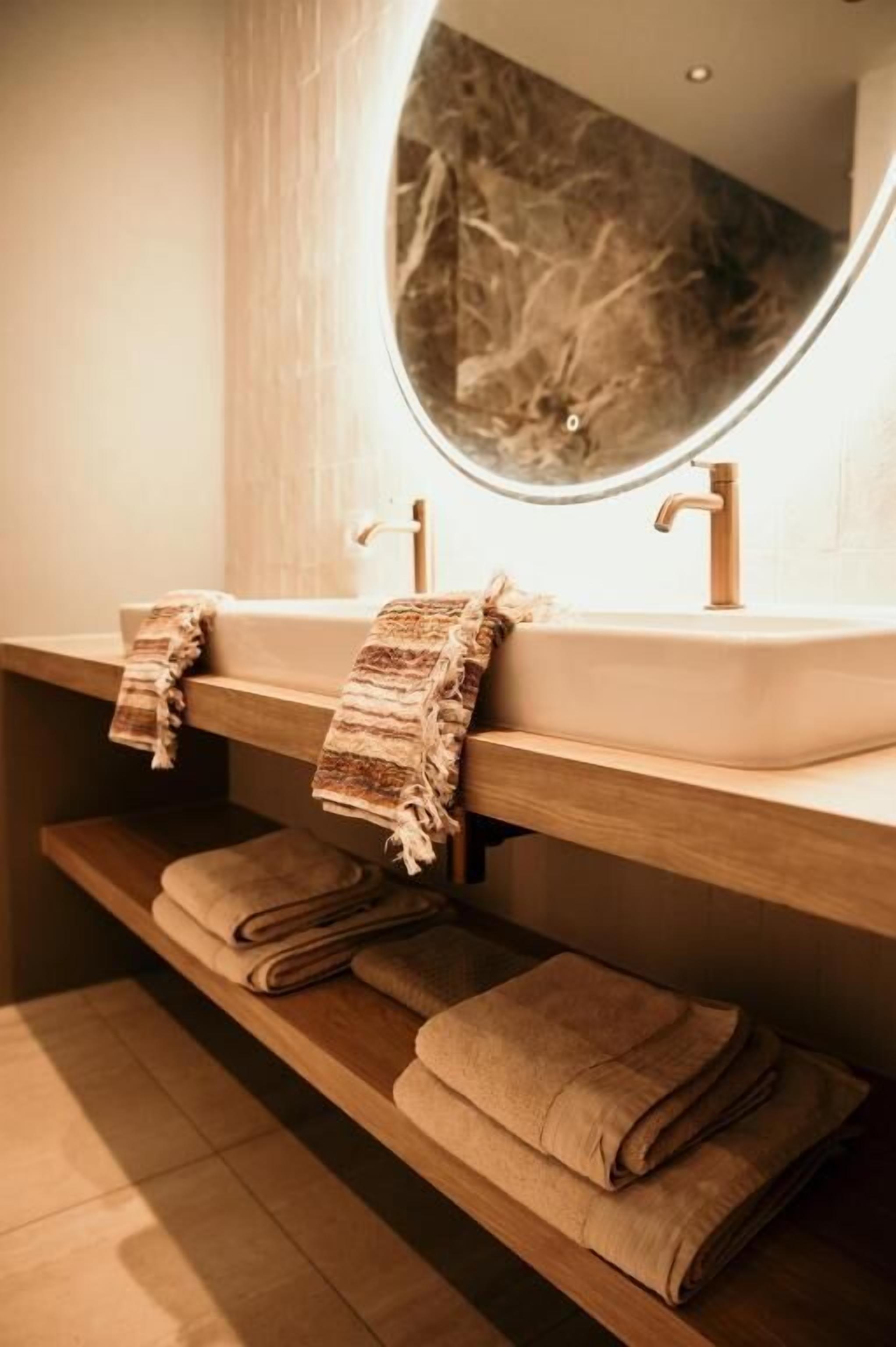 A bathroom with a wooden vanity, a round mirror, and towels neatly folded on the shelf.