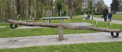 Un sube y baja con extremos metálicos en un parque, con un banco y árboles al fondo.