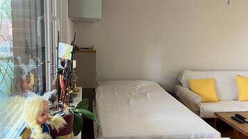A room with a bed, a sofa with yellow cushions, a window with curtains, and a desk with various items including a doll and a vase.