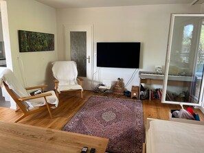 Ein Wohnzimmer mit großem Flachbildfernseher, einem weißen Sessel, einem Holztisch und einem gemusterten Teppich.