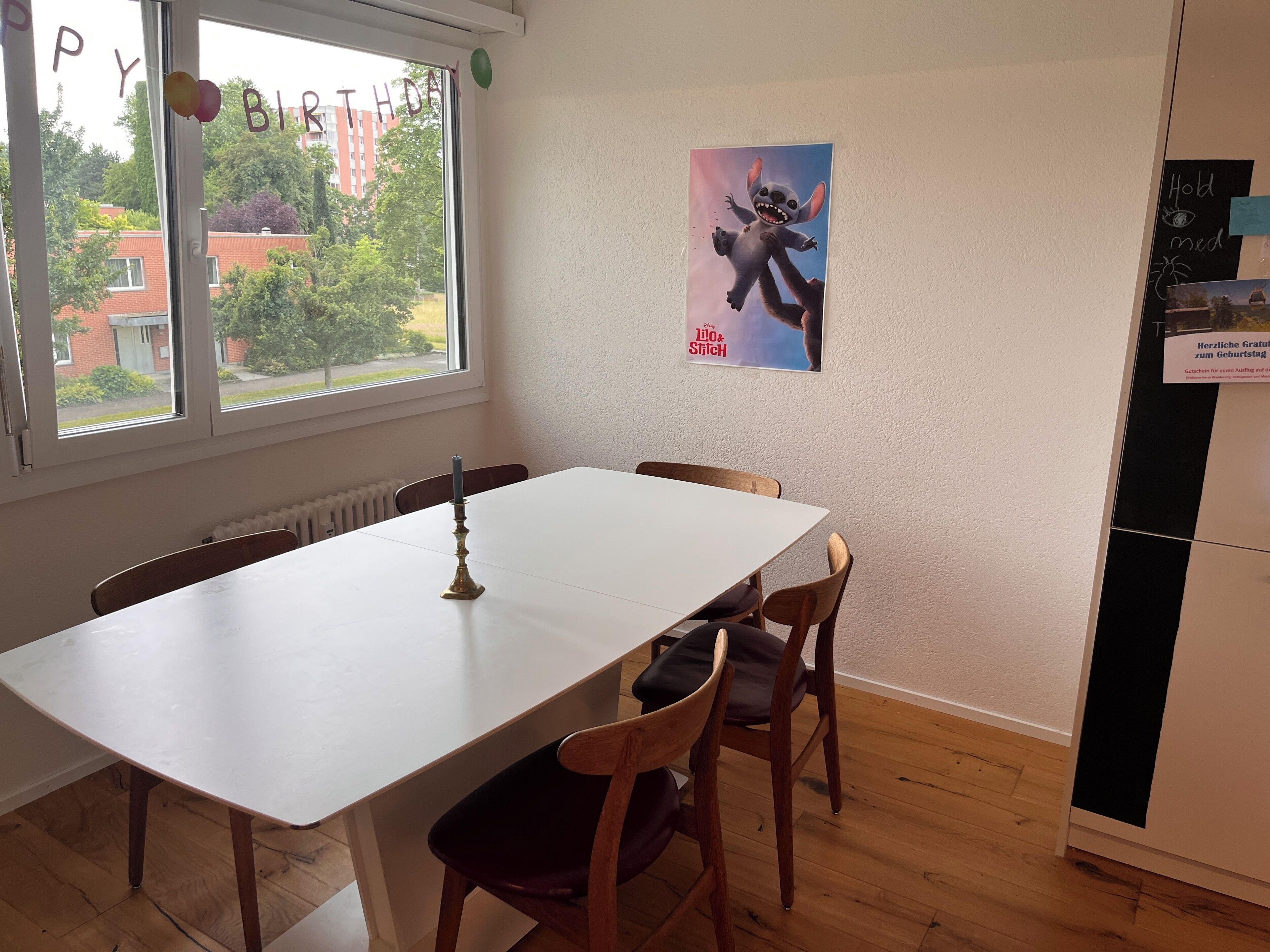 Un coin repas avec une table blanche, des chaises en bois, une fenêtre donnant sur des immeubles et une banderole d’anniversaire.