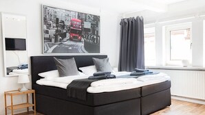 A modern bedroom with a black sofa bed, a flat-screen TV mounted on the wall, a framed cityscape photo, and a window with curtains. - Townhouse Duderstadt: Studio 2 (Duderstadt)