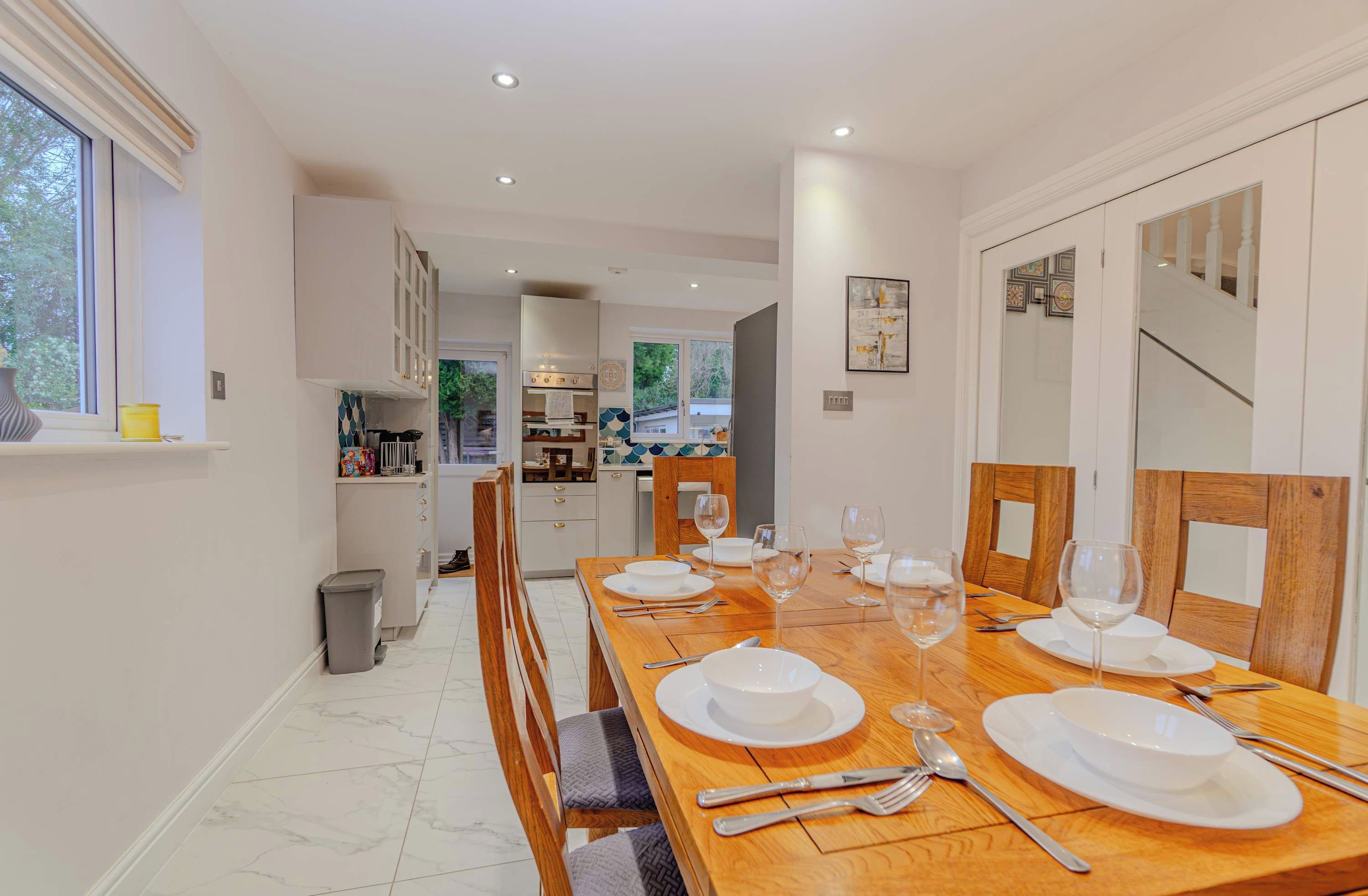 A dining area with a long wooden table set for six, kitchen cabinets, and a view of the adjacent kitchen.