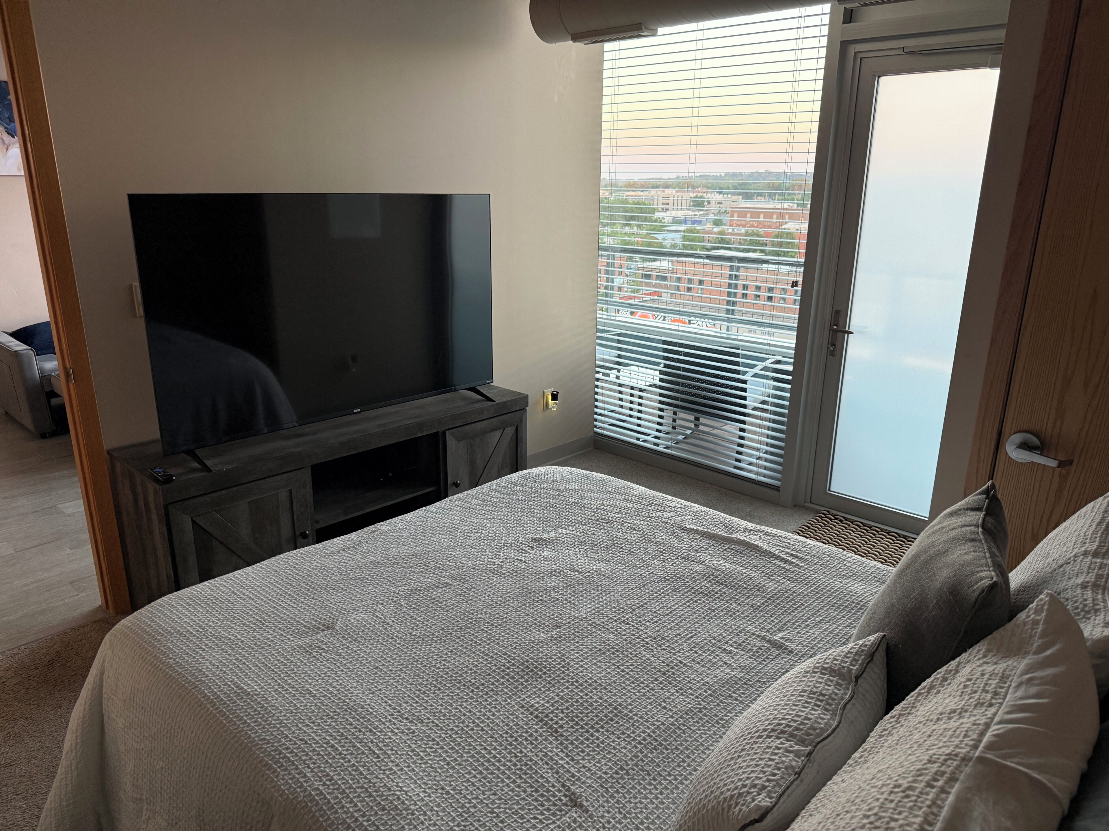 A bedroom with a large flat-screen TV on a wooden stand, a bed with grey bedding, and a view of a cityscape through a window with blinds.