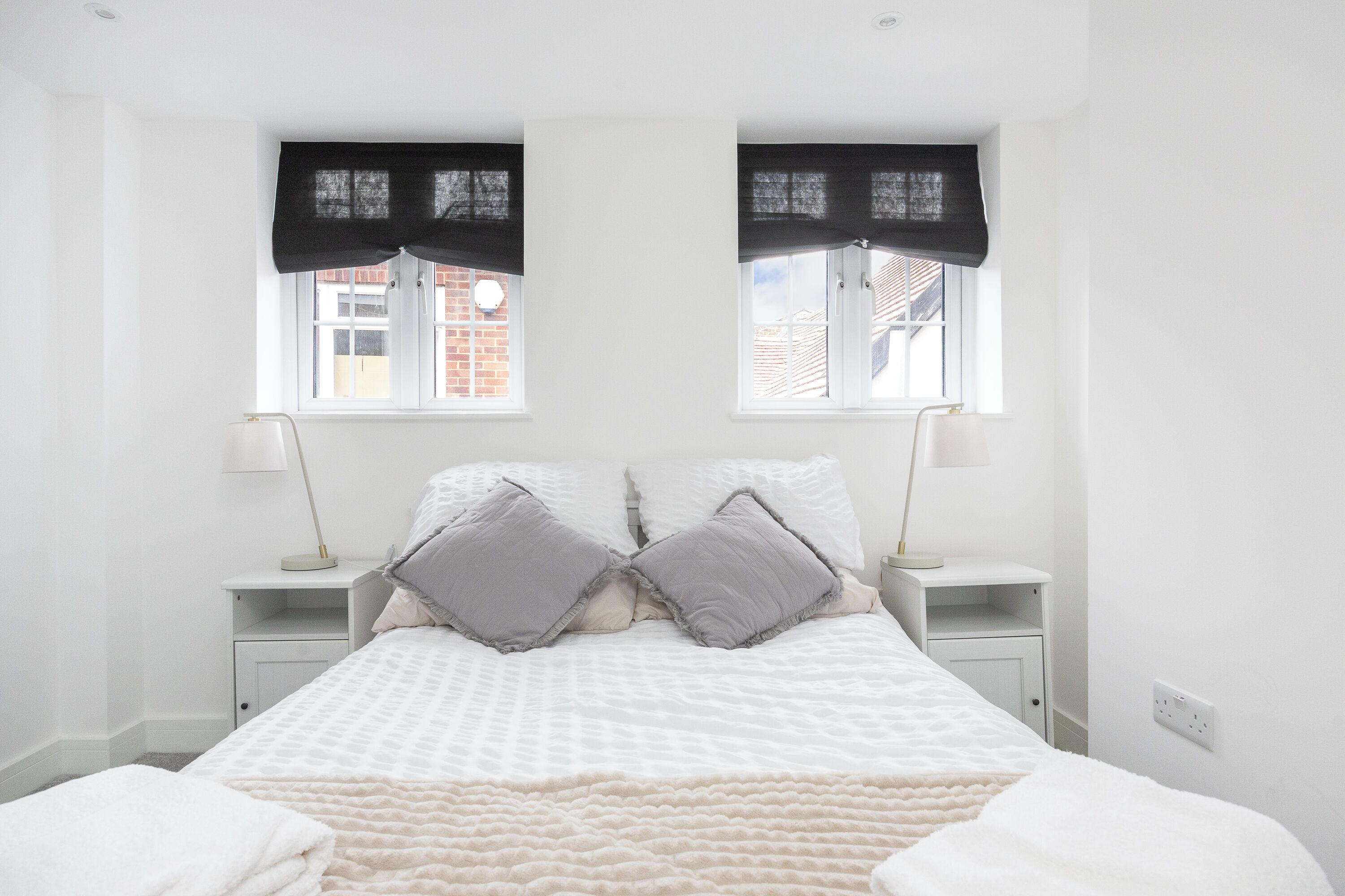 A bedroom with a bed, two bedside tables, two lamps, a window with a view of buildings, and a white wall.
