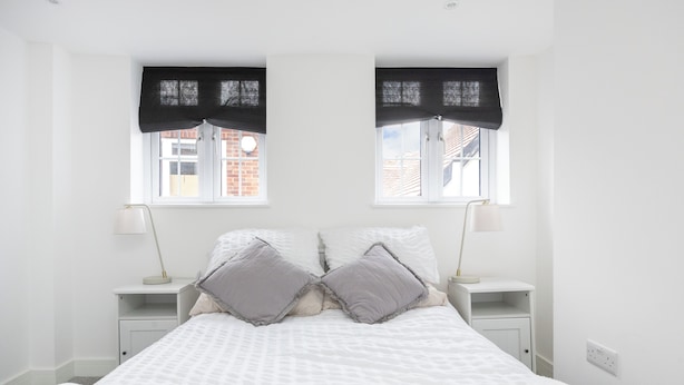 A bedroom with a bed, two bedside tables, two lamps, a window with a view of buildings, and a white wall.