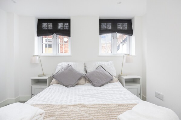 Un dormitorio con cama, dos mesitas de noche, dos lámparas, una ventana con vistas a edificios y una pared blanca.