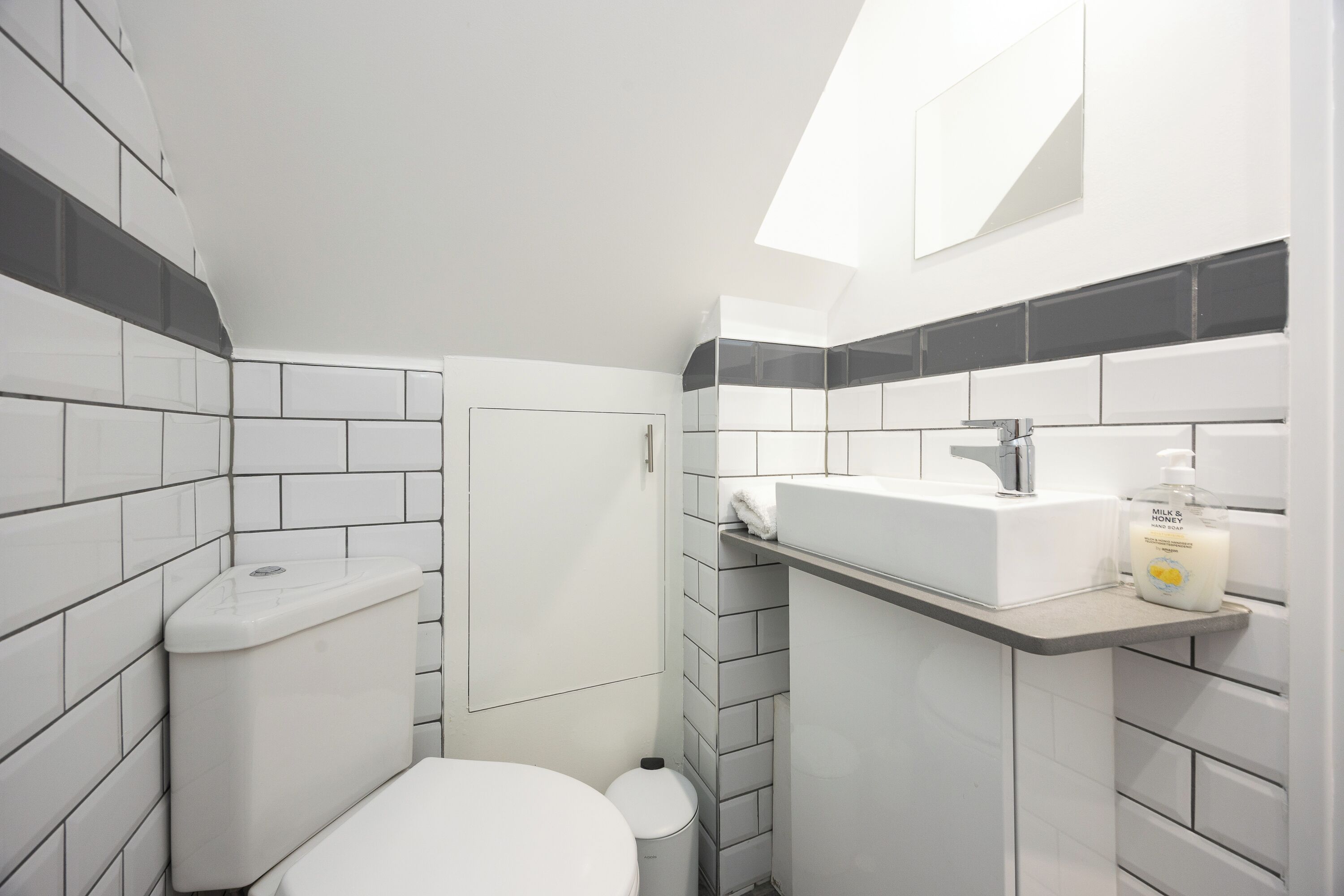 A compact bathroom with white and gray tiles, a white toilet, a sink with a mirror, and a bottle of soap.