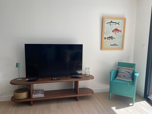 A living room with a flat-screen TV on a wooden stand, a teal armchair with a patterned cushion, and a framed fish painting on the wall. - 1-bedroom apartment (T1), 42 m*, with an 85 m* garden and private parking (Trébeurden)