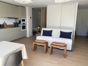 A modern living room with a sofa, two wooden side tables, a dining table with a white tablecloth, and a kitchen area with cabinets and appliances. - 1-bedroom apartment (T1), 42 m*, with an 85 m* garden and private parking (Trébeurden)