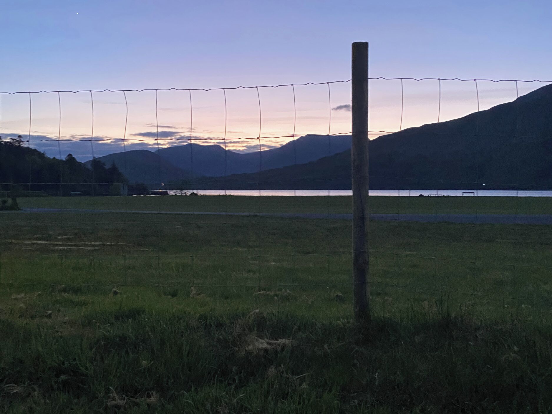 Un champ herbeux avec un poteau en bois et une clôture en fil de fer barbelé, d’où l’on a une vue sur un lac et des montagnes.
