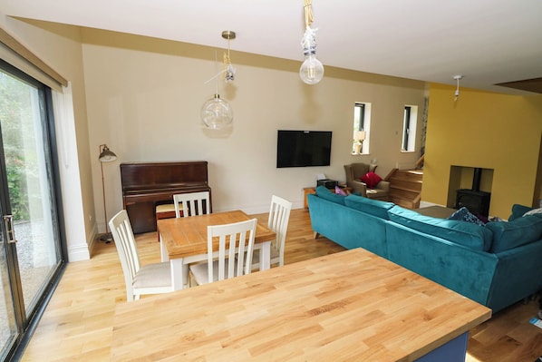 A modern living room with a wooden dining table, white chairs, a teal sofa, and a flat-screen TV mounted on the wall.