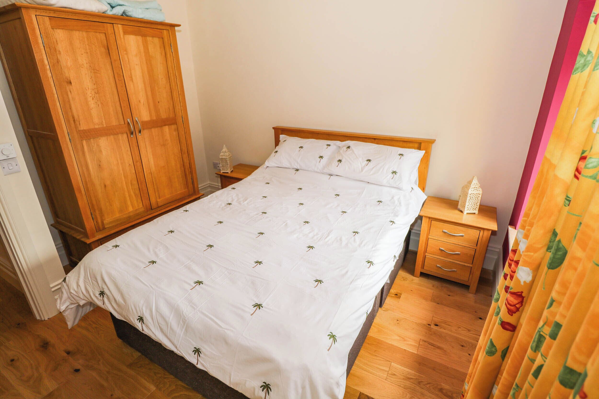Une chambre à coucher avec une armoire en bois, un lit simple avec une literie blanche, une table de chevet et une petite table sur laquelle est posé un objet décoratif.