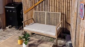 Um banco de madeira com encosto trançado, uma pequena mesa com vasos de plantas e um painel de bambu com placas decorativas.