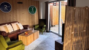 Uma sala de estar moderna com um sofá de couro marrom, uma poltrona verde e uma mesa de centro de madeira. Há duas placas circulares na parede, uma indicando uma escola e outra uma estância de esqui.