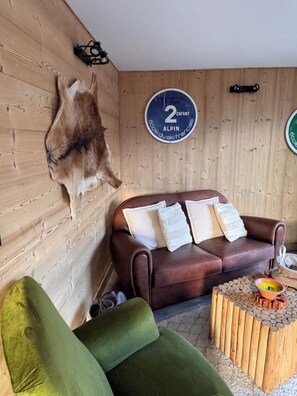 Ein Zimmer mit einem braunen Ledersofa, einem grünen Sessel, einem Holztisch mit einer Schüssel und Besteck, einem aufgehängten Tierfell und einem Wandzeichen.