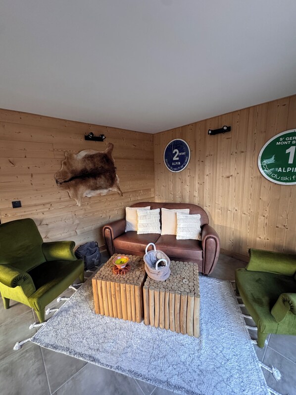 Ein Zimmer mit einer Holzwand, einem braunen Sofa, grünen Sessel und einem Couchtisch mit einem dekorativen Gegenstand sowie einem aufgehängten Tierkopf.