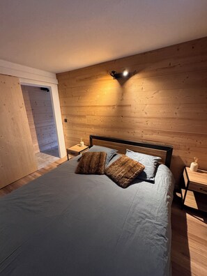 Ein Schlafzimmer mit hölzernem Kopfteil, einem Bett mit grauer Bettwäsche, zwei braunen Kissen, einem Nachttisch mit Lampe und einem Beistelltisch mit Kerze.