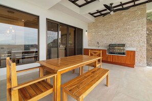 An outdoor kitchen area with a wooden table and benches, a stone wall, and a view through large glass doors.