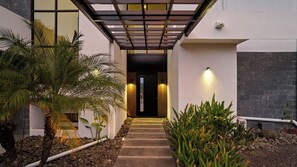 A modern building entrance with a covered walkway, palm trees, and a stone wall.
