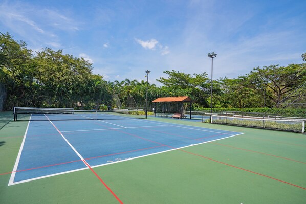 An outdoor tennis court with a net, surrounded by greenery and a gazebo.