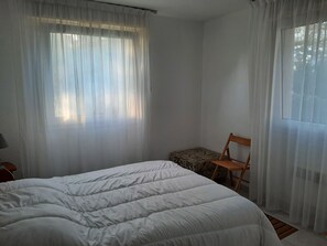 A bedroom with a bed, a chair, and a window with sheer curtains. - Residence with a pool next to the Moliets Golf Course; Duplex apartment for 8–9 people, 10 minutes from the ocean (Moliets-et-Maa)