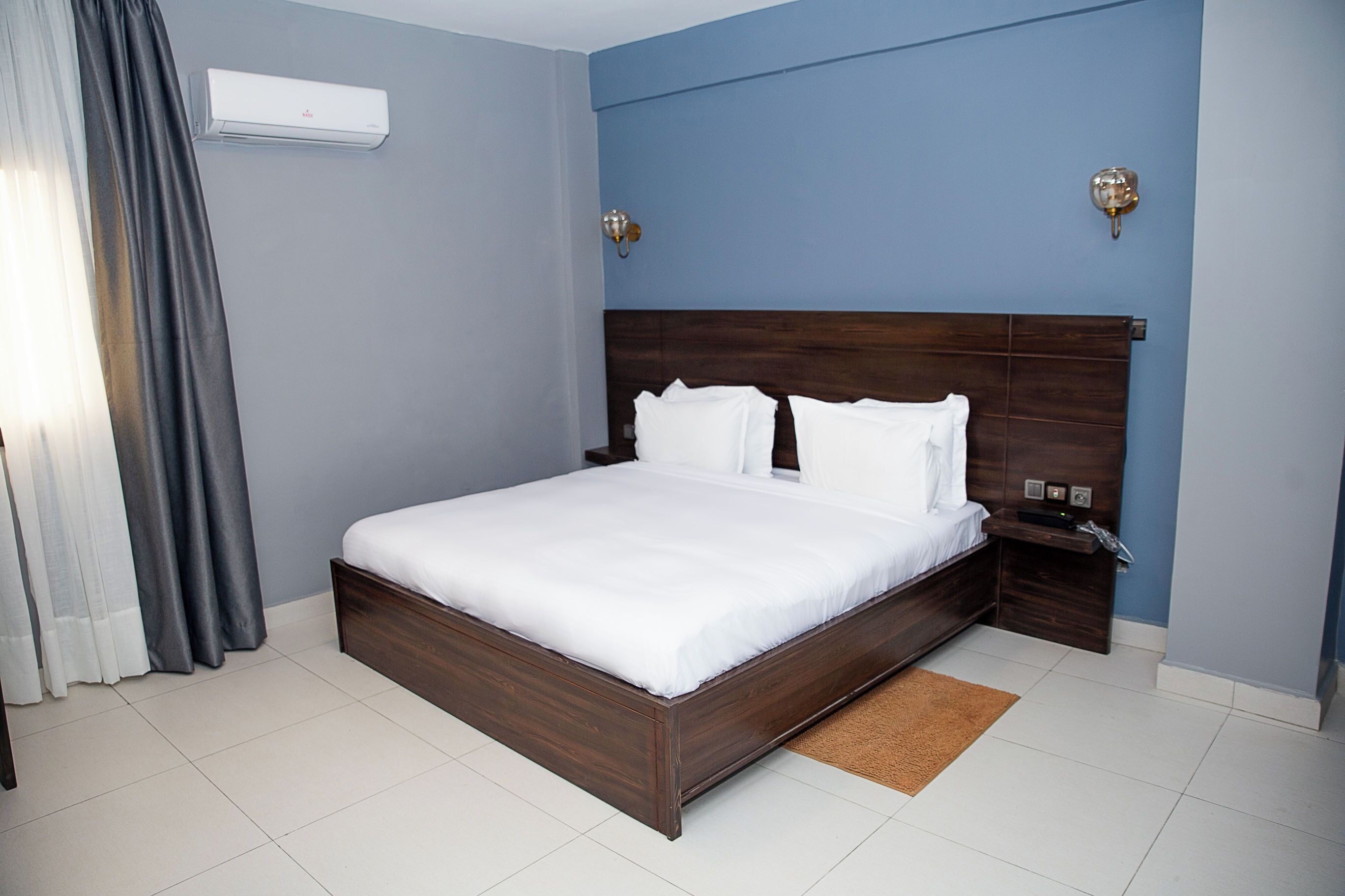 Una habitación de hotel con una cama grande, una mesita de noche, aire acondicionado de pared y una ventana con cortinas.